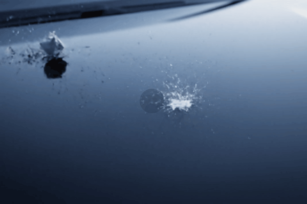 Close-up of a car hood with a fresh rock chip impact, showing cracks and debris from a flying stone.