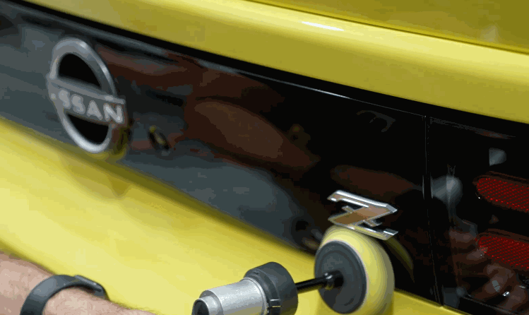 A detailer uses a rotary tool to polish around the emblem of a yellow Nissan Z after ceramic coating application.